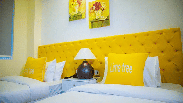 A room with twin beds, yellow cushioned headboard, and framed artwork on the white wall - Lime Tree Hotel Vrindavan