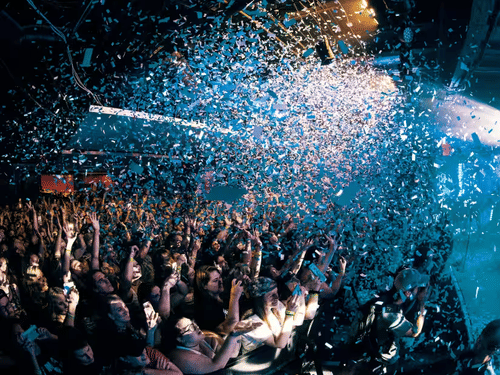 A crowded indoor area with confetti falling and people raising their hands.