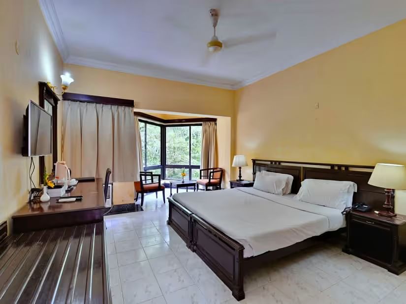 A spacious room with a large bed, a wall-mounted television opposite it, and a full-length window to the side, accompanied by wooden furniture.