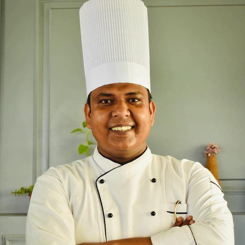 A smiling chef in a white chef's hat and jacket stands in a kitchen setting
