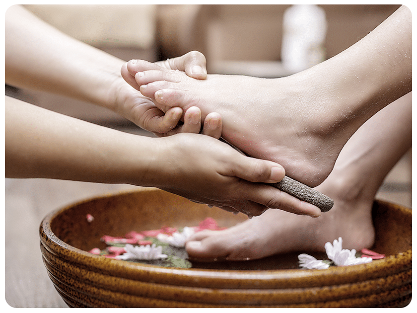 Close-up of a person receiving a relaxing foot massage with a wooden roller over a bowl of water and flower petals.