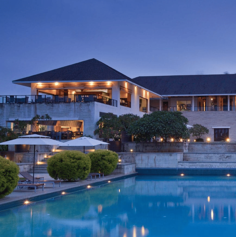 Evening view of The Serai Chikmagalur with illuminated modern villa, steps leading to a pool, poolside loungers, umbrellas, and neatly trimmed greenery.
