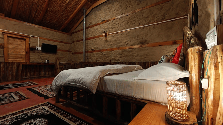 Corner view of the Hamta Peak Cottage featuring a bright table lamp kept on the bed side table along with a wall mounted television in front of it at Amara Upepe - The Sky Village, Manali.
