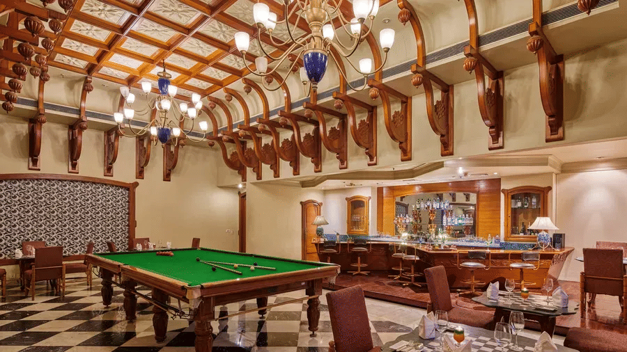 Games and lounge area at The Ummed Jodhpur, Palace Resort and Spa, featuring a central billiards table, classic wooden ceiling beams, checkered flooring and a warm, well-stocked bar counter.