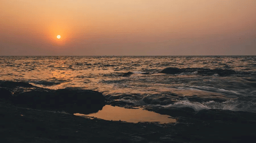Waves gently crash against rocky shore as the sun sets over the horizon, casting reflections on tidal pools.