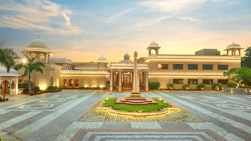 The facade view of Heritage Village Resorts & Spa, Manesar, with driveway, central fountain, and landscaped entrance under a clear sky.