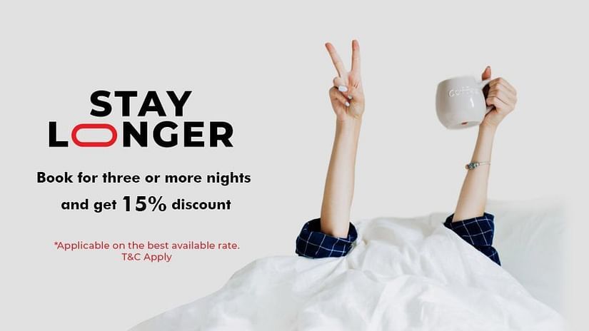 A pair of hands emerge from white bedding, one making a peace sign and the other holding a coffee mug. The text reads: STAY LONGER. Book for three or more nights and get 15% discount. *Applicable on the best available rate. T&C Apply.