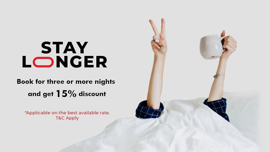A pair of hands emerge from white bedding, one making a peace sign and the other holding a coffee mug. The text reads: STAY LONGER. Book for three or more nights and get 15% discount. *Applicable on the best available rate. T&C Apply.