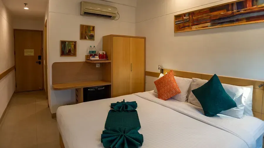  A hotel room with a double bed, neatly made with a dark green folded cloth at the foot. A decorative abstract painting is on the wall, and a wardrobe and a desk with a mini-fridge are on the right side of the image. The room is well-lit and appears tidy. 