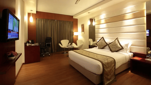 Spacious contemporary hotel room featuring a king size bed with decorative cushions, warm wooden flooring, a work desk, lounge seating area, flat-screen television, and floor-to-ceiling curtains illuminated by ambient lighting.