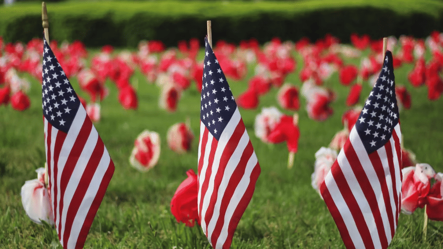 Three small American flags planted in a grassy field with a backdrop of blurred red and white flowers