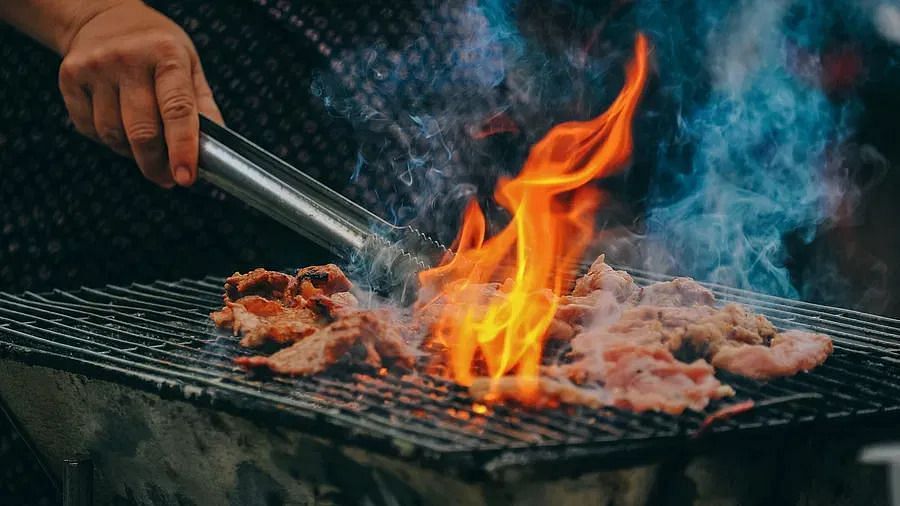 A person using tongs to turn pieces of meat or vegetables cooking on a grill with flames and smoke.