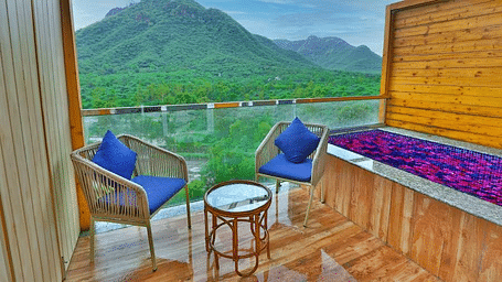 A balcony with two chairs, cushions, and a table overlooks a lush green valley with mountains.- Bamboo Saa Resort & Spa, Udaipur