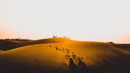 A small group of tourists stand on the peak of a sand dune, with footsteps leading up to them.