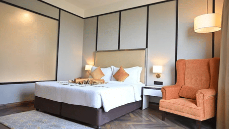 Guest room with king bed, beige paneled headboard, orange pillows and orange wing chair beside pendant lamp at Polo Hotel, Agartala.