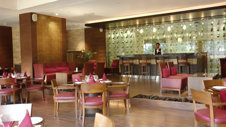 Restaurant dining area at Retreat with wooden chairs and pink upholstered seating arranged around tables with a backlit bar counter and decorative wall panels visible in the background.