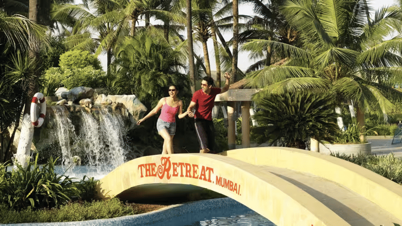 Two people walking on a bridge, surrounded by palm trees and greenery, with a waterfall in the background.