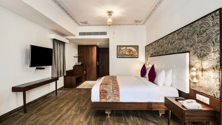 A well-lit hotel room with a plush bed, pillows, a desk, and a TV - Bamboo Saa Resort & Spa, Udaipur.