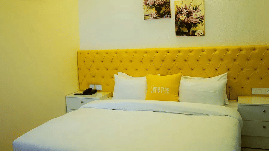 A clean hotel room with a double bed, crisp white linen, a bright yellow panelled headboard, and bedside tables.