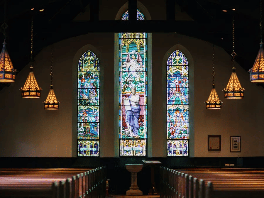 Peaceful church interior with wooden pews and tall stained-glass windows casting coloured light across the aisle.