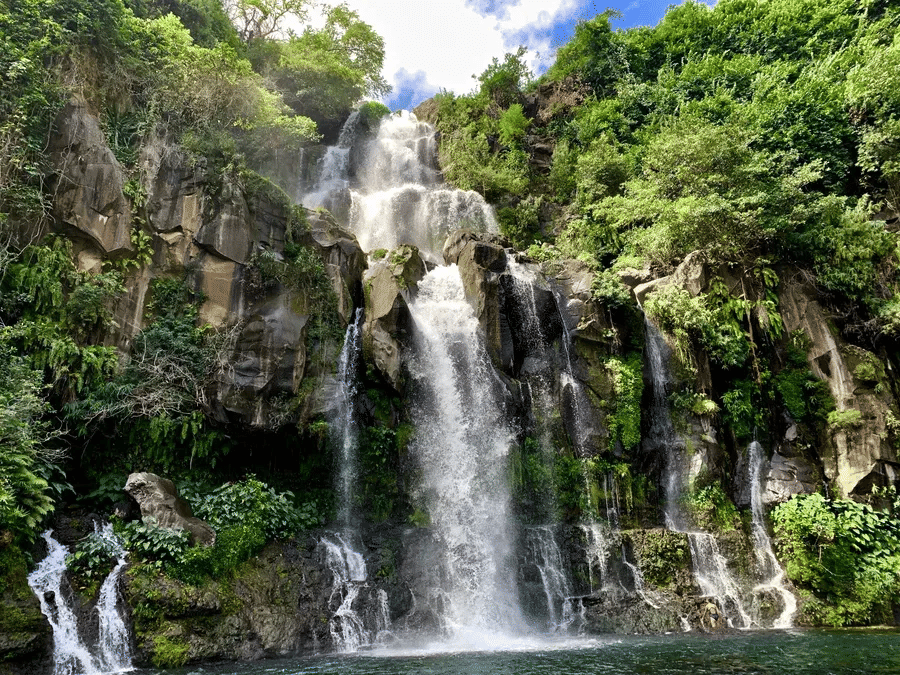 Multi-tiered waterfall cascading through dense green forest, surrounded by moss-covered rocks and tropical foliage under soft daylight.