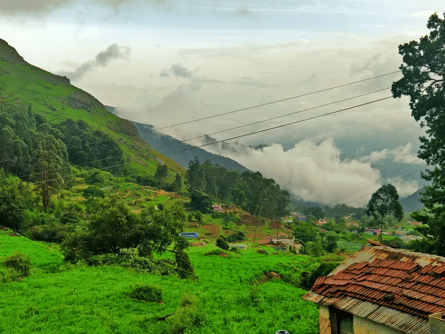 Rolling green hills and mist-covered valley with tiled rooftop in foreground, set against cloudy mountain landscape.