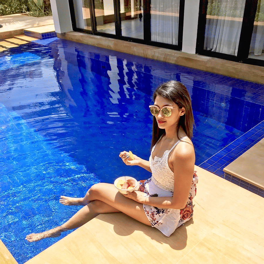 A young woman in a white top sits by the edge of a bright blue swimming pool with her feet in the water.