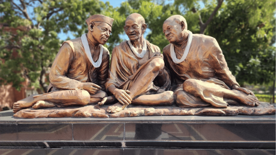 A bronze statue depicting Mahatma Gandhi seated between two other figures, all wearing garlands.