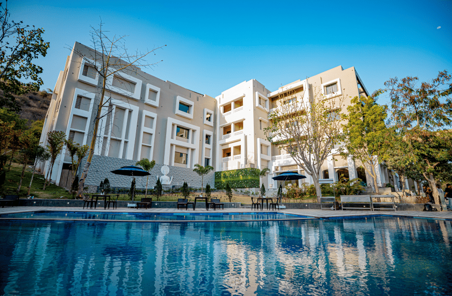 Facade of Essentia Luxury Resort and Spa, Udaipur with the pool in front of it.