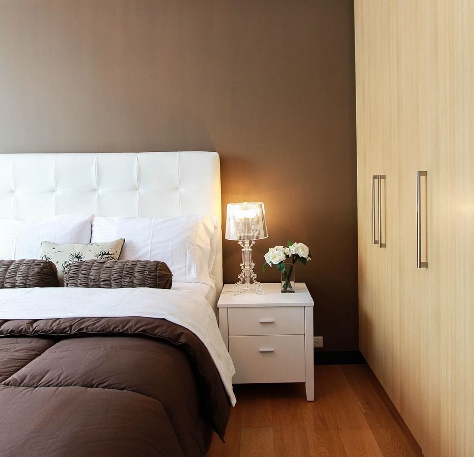 Bedroom with a bed, pillows, blanket, bedside table with lamp and flowers, and a wardrobe with sliding doors.
