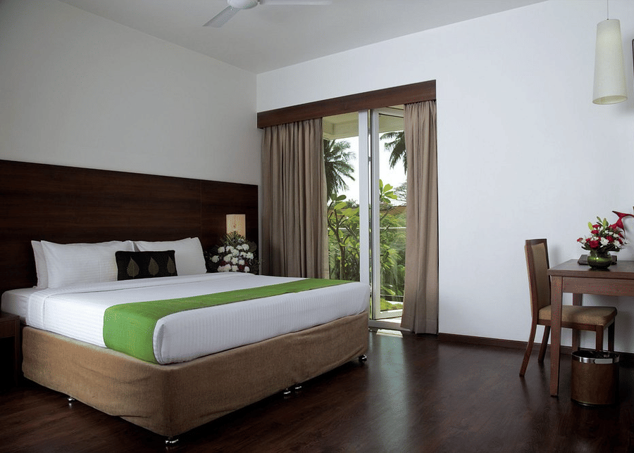 The Garden View Room with a king size bed dressed in crisp sheets and a bright  bed runner. To the right of the bed, there is a small wooden writing desk with a chair, and a large sliding glass door opening up to a balcony with a view of green trees at Tem