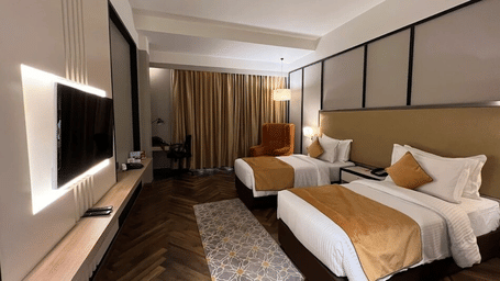 Twin-bed room with backlit TV panel, work desk, orange wing chair, beige curtains and patterned rug at Polo Hotel, Agartala.