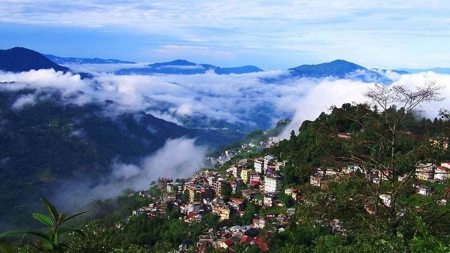  A panoramic view of a hillside town nestled amidst lush green mountains, with clouds swirling below and around the peaks under a blue sky.