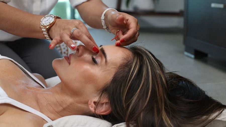 A woman with brown hair lies down, receiving a facial treatment with hands gently touching her forehead.