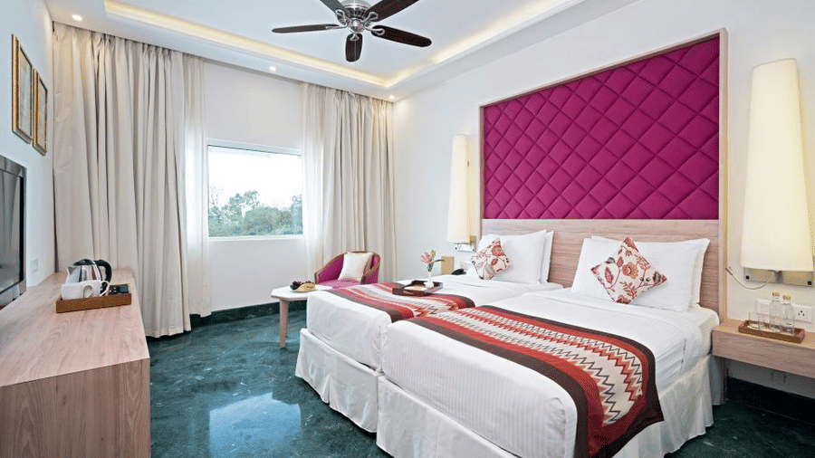 Superior Room at Hotel Clarks Amer, Jaipur featuring twin beds, bedside table with lamp, a chair and centre table, a cabinet table, flat TV screen, and a ceiling fan.
