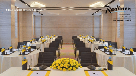 A modern conference room at Radisson Bengaluru with neatly arranged tables, floral centrepieces and meeting materials promoting a special business meeting offer.