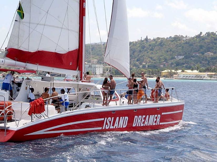 A vibrant red and white catamaran named ISLAND DREAMER sails on blue water, filled with people enjoying the sunny weather.