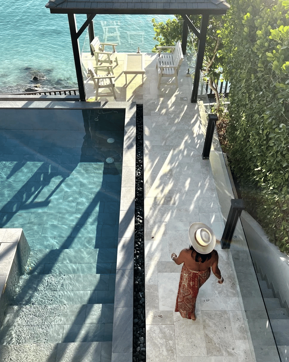 High-angle view of a woman in a wide-brimmed hat and patterned dress walking on a sunny patio next to an inviting pool and ocean.