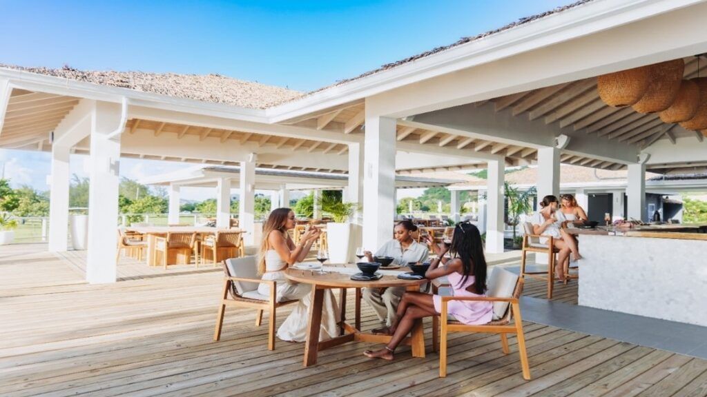 People are seated at tables on a wide wooden deck under a white shaded structure, enjoying drinks and conversation with lush greenery and blue sky in the background.