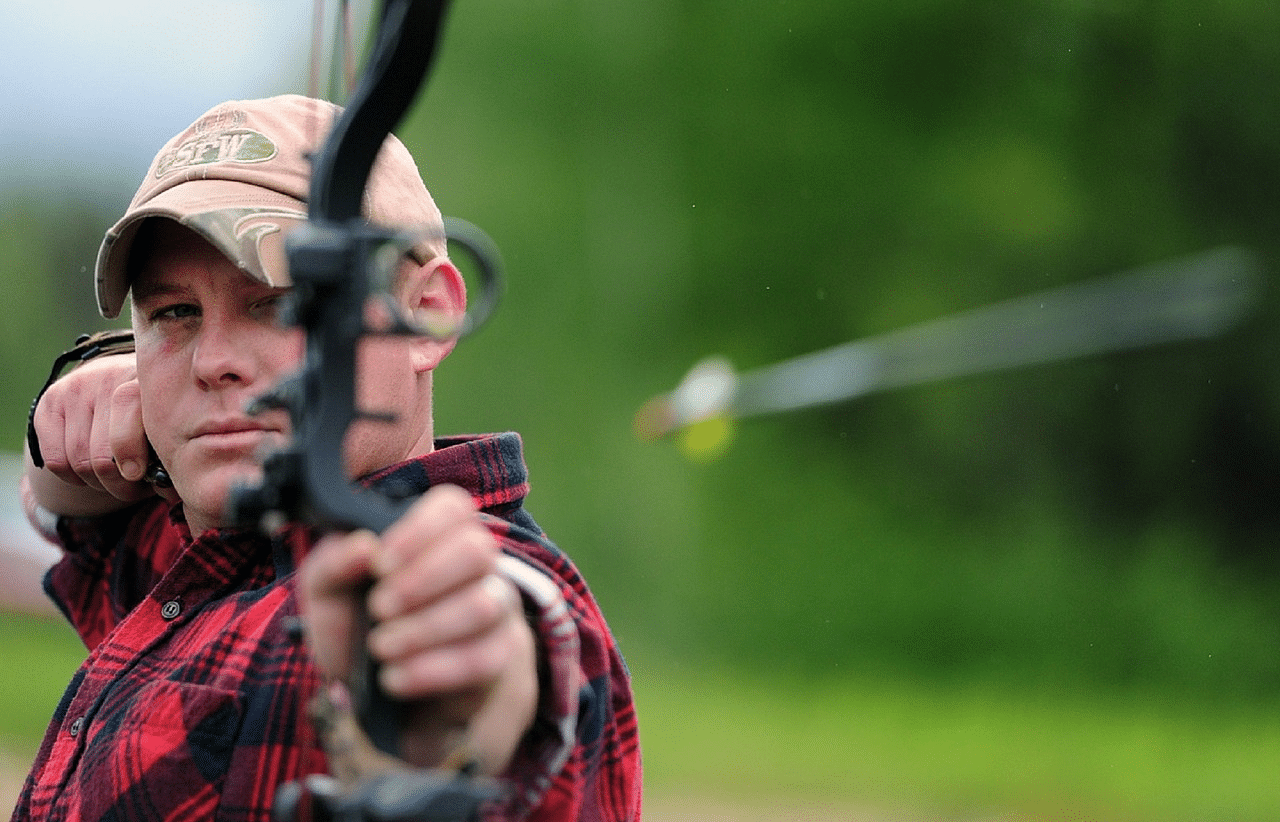 A close-up of a man in the midst of archery, with the arrow just released from the bow.