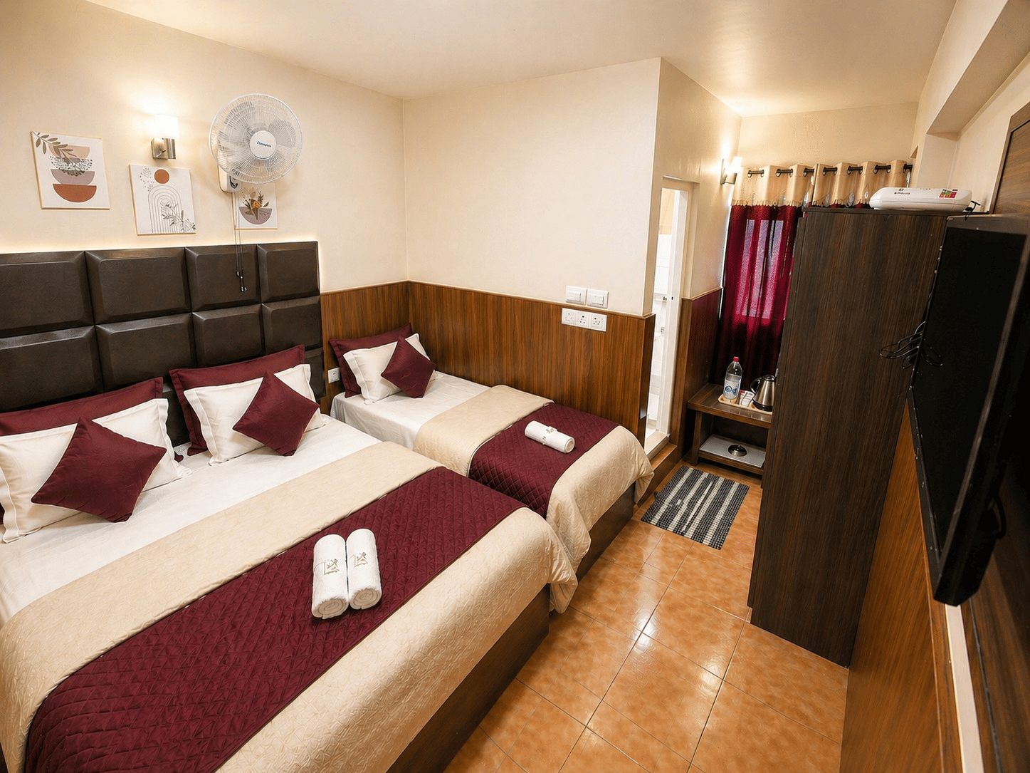 A double bed and a single bed arranged together with folded towels, a wardrobe, and a television in the Deluxe Triple Room at BN Tourist Home, Ooty.