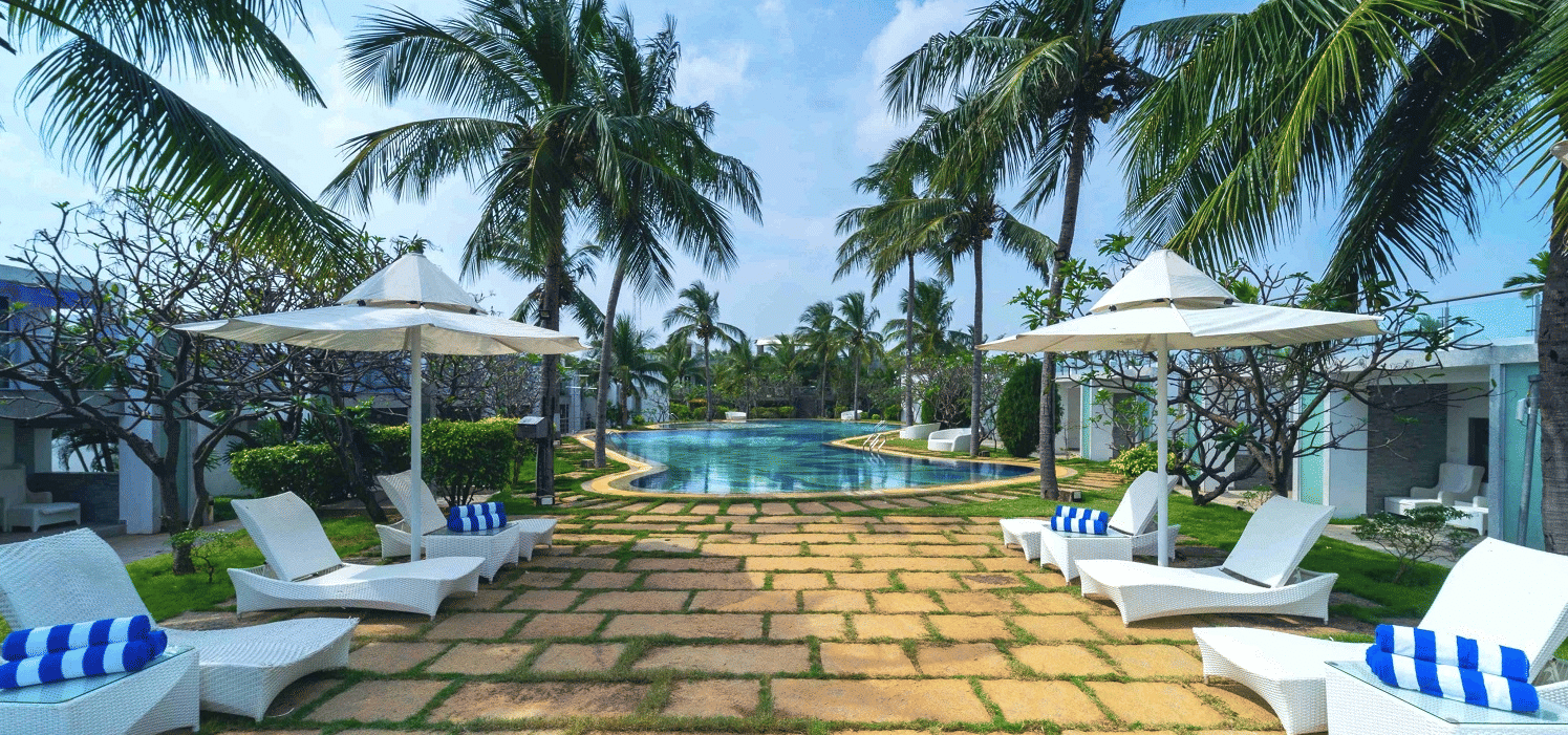 A pathway lined with sun loungers and striped towels leading to the pool, surrounded by coconut trees at Grande Bay Resort & Spa, Mamallapuram.