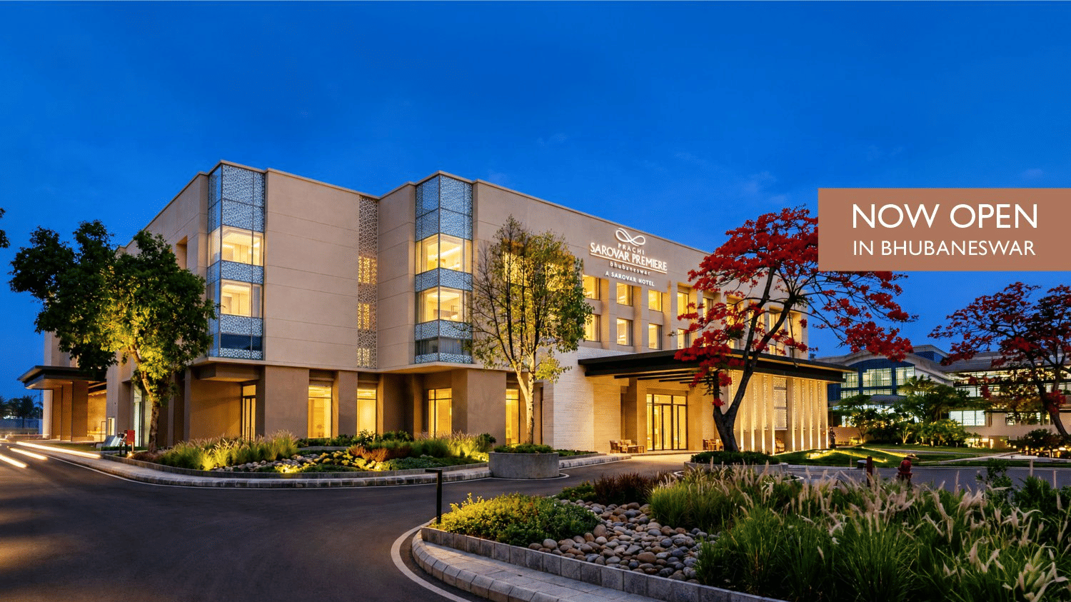 Website banner of Prachi Sarovar Premiere hotel in Bhubaneswar showcasing the brightly-lit facade and driveway of the hotel seen under a dark sky.