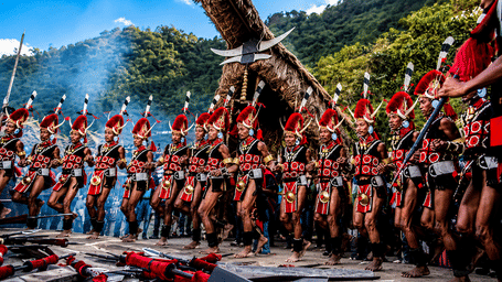 Group of tribal dancers performing in traditional attire and headgear outdoors against a mountain background.