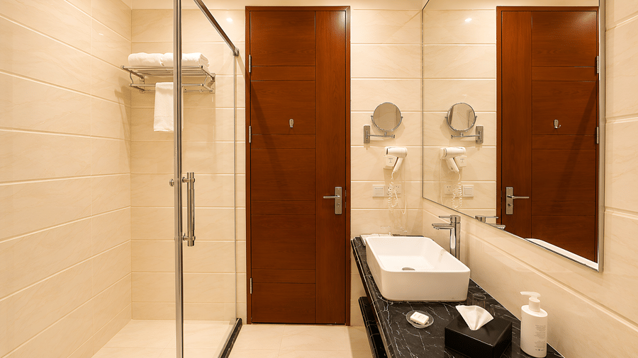Deluxe room bathroom at Golden Tulip Kukas Jaipur featuring a glass shower enclosure, wooden door, wall-mounted mirrors, and modern fittings.