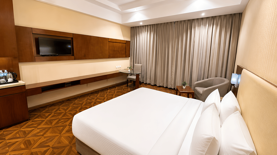 Deluxe room at Golden Tulip Kukas Jaipur showing a king bed with white pillows, wall-mounted TV, patterned carpet, and floor-length grey curtains.