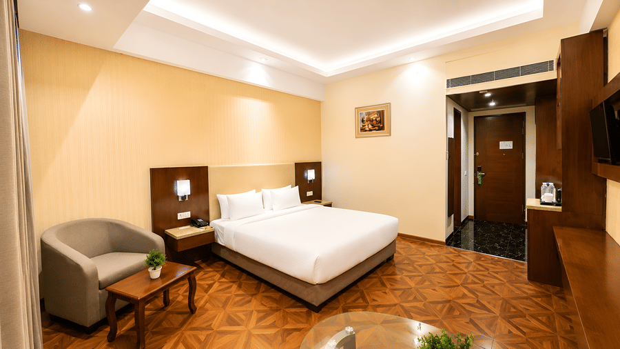 Wide-angle view of the Deluxe room at Golden Tulip Kukas Jaipur featuring a king bed, armchair, wooden flooring, and warm recessed lighting.