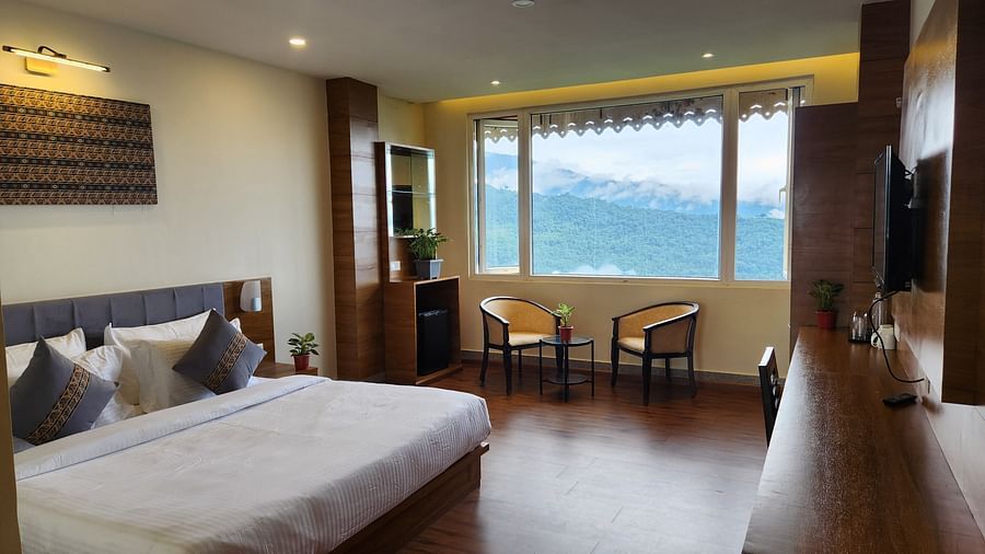 A room that has a bed and two chairs beside a huge window that offers scenic mountain views | Voyage The Royal Retreat, Gangtok