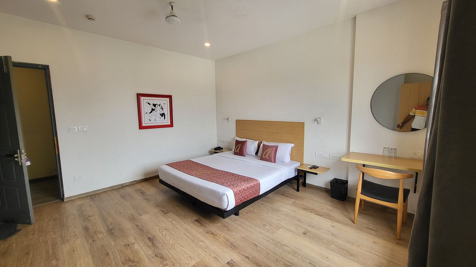 A spacious hotel room with a minimalist design, wooden flooring, a double bed, a desk, and a large circular mirror - Astra Hotels & Suites, Sarjapura