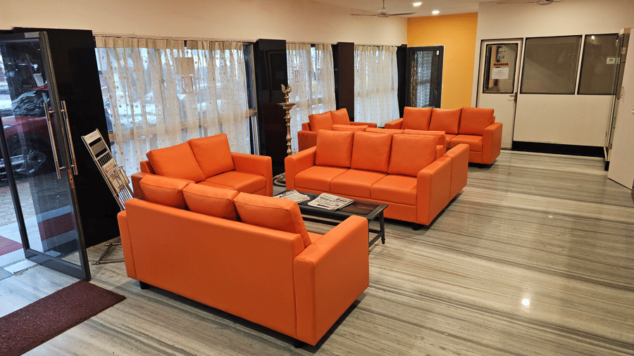 An indoor waiting lounge with several sets of orange couches placed on a light-coloured tiled floor next to large windows with sheer curtains - Hotel Pandian